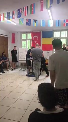 In a room with white-tiled floors and large windows, two men are engaged in an arm wrestling match. The man on the left wears a black shirt and shorts, while his opponent is dressed in a grey shirt and cargo pants. A third man stands between them, seemingly acting as a referee. Various flags hang from the ceiling and a Turkish flag and Romanian flag hang from the wall. A whiteboard with writing is visible in the background. People watch the arm wrestling match.