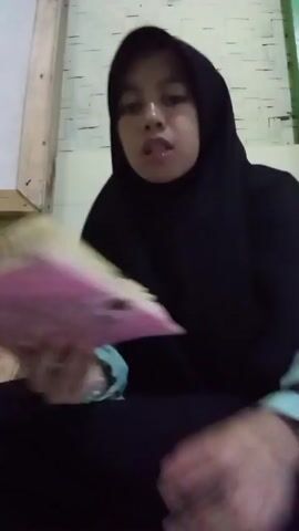 A young woman wearing a black hijab and dark clothing sits on the floor, looking down at an open book in her hands. The book has a pink cover with some green pages visible. She appears to be reading or reciting from the book, as her mouth is slightly open and her head is tilted. Behind her, a textured wall is visible, and to her left, a wooden easel with a white board is propped up. The lighting is dim, with the focus on the woman and the book.