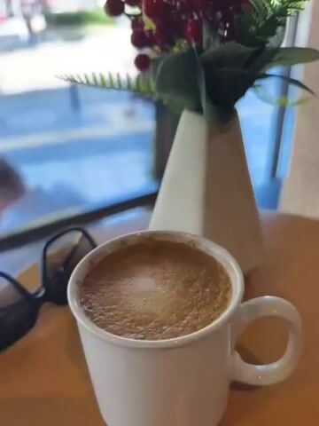 The video shows a close-up of a white mug filled with a frothy coffee drink, placed on a wooden table. Next to the mug are a pair of sunglasses and a small vase containing a faux floral arrangement with red berries. Beyond the table, through a large window, a street scene unfolds.

The street outside appears to be a city environment, possibly a cafe or restaurant setting given the proximity to the street. People are seen walking on the sidewalk and crossing the street. Buildings line the opposite side of the street, and trees provide shade. The overall impression is a bright, daytime scene, likely late morning or early afternoon due to the sunlight casting shadows. The presence of a coffee and the relaxed atmosphere suggest a moment of leisure.

The video pans slightly, revealing more of the street and people. One person is visible sitting at a table inside, looking at their phone, while others are outside. The location cues, such as "İZMİR" on a sign and the general architecture, suggest the city of İzmir, Turkey. There are no explicit indications of the weather other than it appears to be sunny. No specific emotions are overtly conveyed, but the scene evokes a sense of everyday life and a pause for a coffee break.

Visible text includes the word "İZMİR" on a street sign in the background. The overall mood is casual and observant, as if capturing a fleeting moment from a comfortable vantage point.