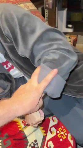 The video shows a close-up view of a person with blonde hair wearing a gray hoodie. The person is hunched over, with their face obscured. The background is a patterned rug with red and cream colors. A hand is visible in the lower left corner of the frame, and another hand with painted fingernails is visible near the person's head. The lighting is dim.