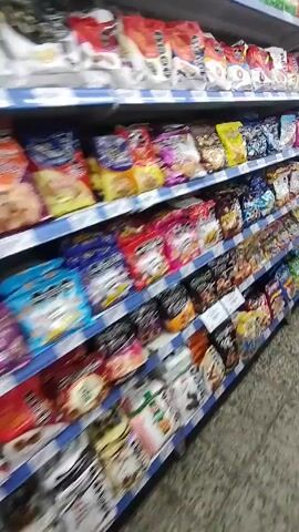 The camera moves along a supermarket aisle with shelves stocked with bags of cookies. The shelves are full from top to bottom, showcasing a wide variety of brands and flavors. The floor is tiled in a light color with darker flecks. The movement of the camera suggests someone is walking or pushing a cart along the aisle.