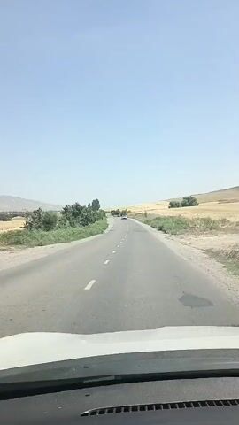 A car's dashboard is visible at the bottom of the frame, facing forward. Beyond the windshield, a two-lane asphalt road stretches into the distance under a clear, bright blue sky. The road has white dashed lines marking the lanes. To the left of the road, there are trees and green vegetation. To the right, there are fields of dry grass and some scattered bushes, with gently rolling hills in the background. A few other vehicles are visible on the road ahead, appearing as small white dots. The overall scene depicts a daytime drive through a rural, possibly dry, landscape.