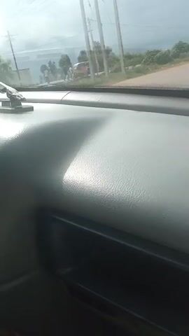 The video is shot from inside a vehicle, showing the dashboard and the view through the windshield. The road ahead is visible, with various vehicles including cars, a van, and a three-wheeled auto-rickshaw. There are also utility poles and some buildings in the background. The sky is overcast. The video appears to be a short clip of a journey.