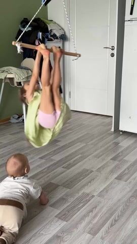 In an indoor setting, a young girl in a light green dress is hanging upside down from a trapeze bar, while a baby in a white shirt and beige pants crawls on the floor beneath her. The girl's brown ponytail hangs towards the floor as she does the trick. The trapeze bar is suspended from the ceiling by ropes. The floor is a light grey wood-look laminate. In the background, there is a white door, a green wall, and a white stool with baby gear on it. There is also a shelf with clothing and other items stacked on it. The girl loses her grip and drops lightly to the ground, standing and turning to face the baby.