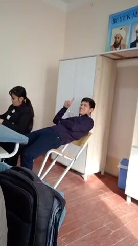 The video shows a classroom setting with two students, a cabinet, and a poster of mathematicians. One student sits at a desk on the left, appearing to be writing or reading. The other student leans back in a chair, his legs stretched out, pointing a finger upwards. The cabinet is white and beige, and a poster of mathematicians is on top of it. The room has an older aesthetic, with wooden floors and simple furniture.