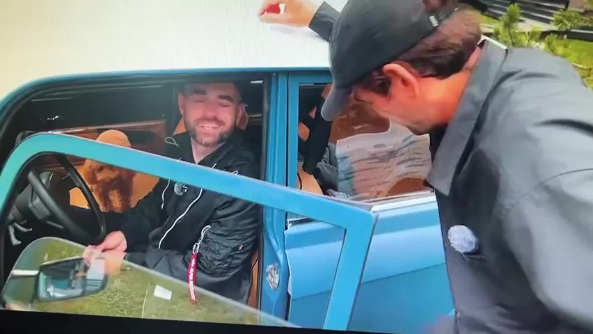A medium shot shows a man with a gray beard and a black cap sitting in the driver's seat of a teal-colored car. He is smiling. A fluffy orange dog is standing on its hind legs behind the steering wheel, with its paws on the wheel, as if driving. The car door is open, and another person, wearing a black jacket and a black cap, is leaning into the car from the right side, partially visible from the back. The car is parked on a grassy area, with trees and foliage visible in the background. The car is a vintage model, possibly a sedan, with a white roof and chrome accents. The license plate is visible, and appears to be Russian.