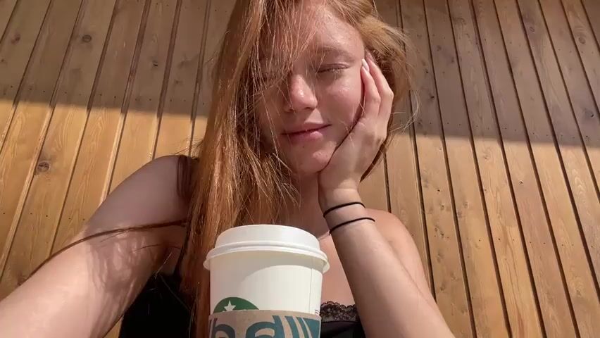 A young woman with long, reddish-blonde hair rests her chin on her hand and looks directly at the camera. She is wearing a black top with lace trim. In front of her is a white Starbucks coffee cup with a brown sleeve. The background consists of a wooden wall with vertical planks. The lighting suggests it is daytime, with sunlight casting shadows.