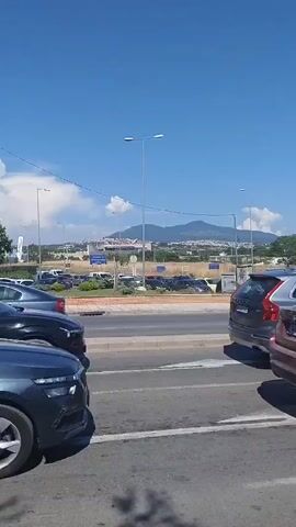 A vehicular traffic scene is depicted under a clear blue sky. The camera pans from right to left, capturing various cars on a road. In the foreground, a red BMW X6 is visible, its rear section and wheel prominently featured. Adjacent to it, a white vehicle, possibly a crossover SUV, is seen. Further down the road, a silver car, then a gray car, and a black car are observed. Traffic lights are present, with one appearing to be red. Trees and distant mountains form the backdrop of the scene. The overall environment suggests an urban or suburban setting.