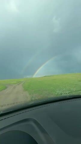 The video shows a view from inside a vehicle looking out at a double rainbow over a green, grassy field. The sky is overcast and grey. A dirt road curves through the field towards the rainbow. There are some white poles or structures visible in the distance on the field. The perspective suggests the vehicle is moving forward.