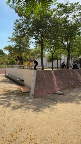 A person wearing a black t-shirt and black pants performs a skateboard trick on a metal rail. The skateboarder grinds on the rail for a moment, then jumps off the rail and onto a mat on the ground. The skateboard lands separately on the ground. The skateboarder runs off to the right of the frame. In the background, trees and buildings are visible. Two other people are sitting in the background watching. The ground is covered in dirt and fallen leaves. The sky is blue.