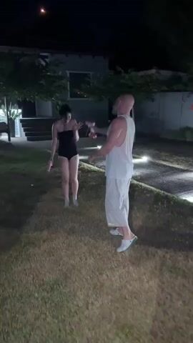 In this video, two individuals are engaged in a water gun fight on a grassy lawn at night, likely in Nea Potidea, Greece. The scene is illuminated by soft, warm outdoor lighting, suggesting a pleasant evening.

The subjects are a woman in a black swimsuit and a man in a white tank top and light-colored pants. Both are holding water guns. The woman appears to be reacting to being shot with water, with her hands up in a defensive posture, and she is also holding a water gun. The man is actively aiming his water gun towards the woman, spraying water at her.

The setting is a backyard with a well-maintained lawn and a paved pathway with embedded lights. In the background, there is a house with some trees and foliage. The overall atmosphere suggests a playful and lighthearted activity taking place outdoors. The time of day is clearly night, indicated by the darkness and the artificial lighting. No visible text is present in the video. The emotion conveyed is one of fun and friendly competition.