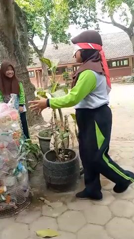 The video begins with a person wearing a brown hijab and a red bandana over their eyes, facing a large cylindrical structure filled with plastic bottles. This person is wearing a gray shirt with bright green accents on the sleeves and a black tracksuit with green stripes. They have their hands extended as if reaching for something. To the left of the bottle structure, another person wearing a brown hijab and a bright green shirt is visible, smiling and clapping their hands. The environment appears to be outdoors, with trees and buildings in the background, suggesting a school or public area. The ground is paved and there are plants in pots. The person with the blindfold takes a step to their right, their body twisting as they move. The other person continues to smile and clap. The blindfolded person then turns further to their right, their arms continuing to move. The video then shows the blindfolded person in profile, still moving their arms. The camera pans slightly to the right, showing more of the surrounding greenery and plants. The blindfolded person takes another step, their body is partially obscured by the tree trunk on the left. The other person is no longer visible in the frame. The video captures a short segment of an outdoor activity, possibly a game, where one person is blindfolded and guided or interacting with a structure made of recycled materials.