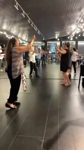 Several people are dancing in a line inside what appears to be a large restaurant or event space. They are facing the same direction and hold their arms up as they move. The dancers are a mix of ages and are dressed in casual clothing. Some are wearing white shirts and black pants, while others wear dresses or patterned tops. There is a band playing in the background on the left. The space has a dark ceiling with multiple light fixtures. The floor is shiny and dark. People are seated at tables in the background, watching the dancers.