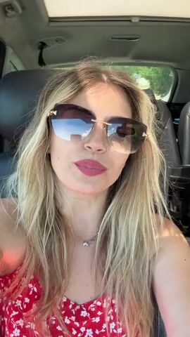 A blonde woman is seated in the passenger seat of a car during daylight hours. She has long, wavy hair and is wearing large, rimless sunglasses with gold accents, which reflect the bright sky and green trees outside. Her lips are painted with red lipstick, and she is wearing a red spaghetti strap top with a white floral pattern. A delicate silver necklace with a small pendant is visible around her neck, and she has several beaded bracelets on her left wrist.

The car's interior features black leather seats, and a sunroof is visible above her head. Through the car windows and reflected in her sunglasses, green foliage and trees can be seen, indicating an outdoor setting, possibly rural or suburban.

The woman is actively singing along to music, moving her lips in sync with the audible song. She smiles frequently, particularly when looking towards the camera, conveying a joyful and relaxed demeanor. She occasionally makes small, expressive hand gestures with her right hand while singing. The scene suggests she is enjoying a car ride on a sunny day.