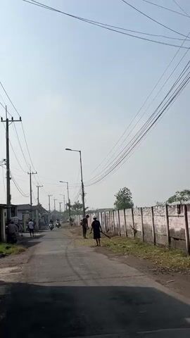 The video shows a paved road in a rural or suburban area. There are houses on the left side of the road and a wall on the right side. Several utility poles with power lines run along the road. The sky is clear and bright. On the road, there are a few people and motorcycles. Two men are on the right side of the road, and one of them appears to be sweeping or raking the ground with a broom or rake. There are also a few motorcycles further down the road, some with riders. The overall impression is a quiet, everyday scene in a residential area.