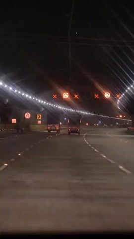 The video shows a car's perspective driving through a tunnel at night. The tunnel is illuminated by rows of white lights along the ceiling and orange lights with red cross symbols, indicating traffic management. Several cars are visible ahead, driving in the same direction. Road signs, including a speed limit of 60 and a curve warning, are visible on the left side of the tunnel. The road surface is asphalt with white lane markings and reflective markers. The camera appears to be mounted inside the vehicle, looking forward.