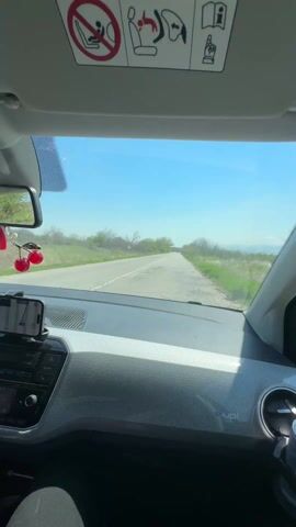The view from the driver’s seat of a car shows it traveling along a straight, asphalt road through a rural area on a clear, sunny day. The road has a white line marking the center, with green vegetation and trees lining both sides. The car's dashboard is a textured light gray with a black infotainment system. A phone is mounted to the dashboard. A Red Bull can is in the cup holder. The car's sun visor is down, displaying a warning label about airbags. Music is playing in the background.
