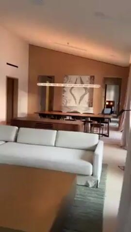 The video starts inside a modern, well-lit room. It contains a large white sofa, a wooden coffee table on a green rug, a long wooden table with chairs underneath a rectangular light fixture, and a piece of art on the wall. The camera moves towards a curtain and then reveals a long balcony with yellow walls and a wooden ceiling. There are several small wooden tables with chairs on the balcony. The video ends with a panoramic view of a calm lake surrounded by mountains under a cloudy sky.