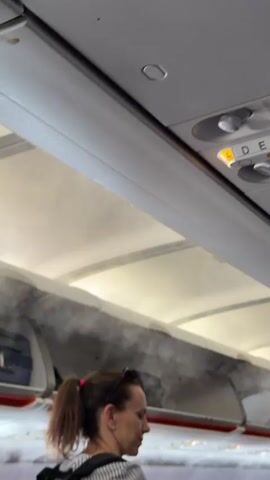 The video shows the interior of an airplane cabin. In the foreground, a woman with her hair in a ponytail is looking to the right. Behind her, a man with sunglasses is partially visible. Above them, overhead compartments are open, and a white mist or vapor is emanating from them, obscuring the view into the compartments. Luggage is visible in one of the open compartments on the left. The ceiling of the plane with lighting and air vents is also visible.