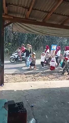A group of children and adults are walking down a paved road. The children are wearing various costumes, including a military uniform and what appears to be traditional attire. Some of the adults are wearing hijabs and white shirts. The group is holding Indonesian flags and a banner with text. The scene is outdoors, with trees and vegetation visible in the background. A motorcycle is parked on the left side of the road. The ground in the foreground is covered with gravel. The overall impression is of a parade or a procession.