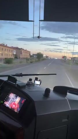 The video captures a first-person perspective from the driver's seat of a bus. The driver, wearing a light-colored shirt and beige pants, is visible on the left, with their hands on the steering wheel. The bus is in motion, driving down a road lined with apartment buildings. The screen in front of the driver displays an image of a person.

The scene is set in Ботақара, Kazakhstan, and appears to be during dusk or early evening. The sky is cloudy with a soft, diffused light, suggesting the sun has recently set or is about to rise. The apartment buildings are several stories high and have a consistent architectural style, with light-colored facades and rows of windows. There are streetlights visible in the distance, indicating a populated area. The road is paved, and there is some vegetation along the sides.

Notable details include the dashboard of the bus, which is filled with various buttons and displays. The YUTONG logo is visible at the top of the windshield, indicating the bus manufacturer. The overall mood is one of a journey concluding or beginning at twilight. The video provides a glimpse into the experience of driving through a Kazakh town as daylight fades.