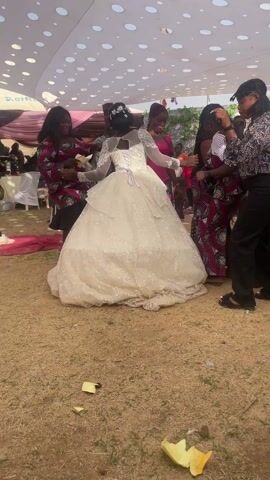 A bride wearing a white wedding dress dances with a group of women on a dirt ground covered by a large, white tent with circular designs. Some women wear vibrant African print dresses, while others are in casual pants and tops. The bride is the center of attention, being surrounded and held by the women as they move to the music. In the background, chairs and tables suggest a wedding reception setting. A few people can be seen further in the background, but the focus remains on the bride and her companions.