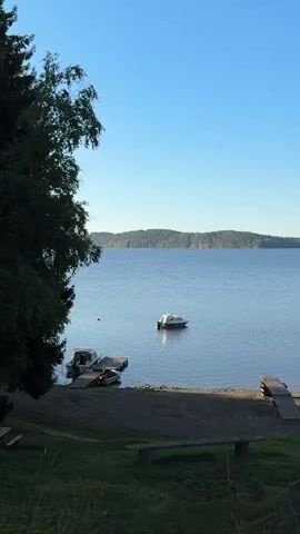 The video pans across a serene lake scene on a clear day. In the foreground, a grassy embankment leads down to a gravelly shoreline. A wooden bench sits on the grass. There are two small wooden docks extending into the calm, blue water. Moored to the docks are several small boats, including a white speedboat and a smaller, dark-colored boat. Further out on the lake, a single white boat is anchored. On the shore, a small, dark-colored wooden structure with a gabled roof sits to the right of the docks, and behind it is a pile of cut logs. To the far right, part of a larger building with a dark roof is visible. The lake extends to the horizon, where rolling, tree-covered hills form the distant landscape under a clear blue sky. A large tree with dark green foliage frames the left side of the image.