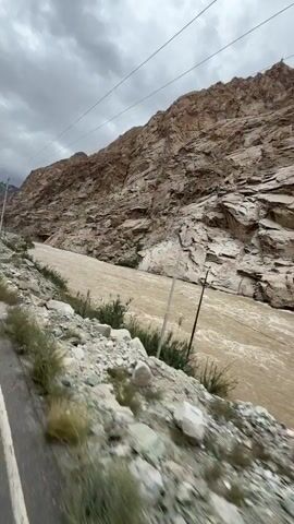 A video captures a muddy, fast-flowing river in a rocky, mountainous area. The river appears swollen and turbulent, suggesting recent heavy rainfall or snowmelt. The terrain is barren and rugged, with steep, rocky slopes on one side and a road or path on the other. There are scattered rocks and sparse vegetation along the riverbank. Overhead power lines are visible against a cloudy sky. The video appears to be shot from a moving vehicle, giving a sense of travel through this dramatic landscape.