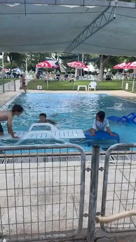 The video shows an outdoor swimming pool with a group of children playing. In the foreground, a metal fence with a concrete base lines the pool. Three children are in the water. One child on the left, wearing green patterned shorts, is pushing a white floatation device. Another child in the middle, wearing a black swimsuit, is also near the float. A third child on the right, wearing a light blue shirt, is sitting on a blue float. The water is clear and blue. In the background, there are lounge chairs, umbrellas with red and white patterns, trees, and more people. The pool area is covered by a white canopy.