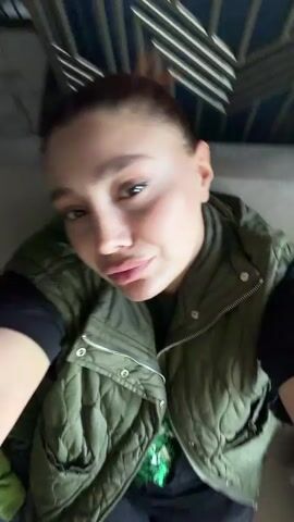 The video shows a woman with brown hair pulled back, wearing a green quilted vest over a black shirt, and ripped jeans. She is seated and looks up at the camera. The background appears to be a building interior with a decorative ceiling.