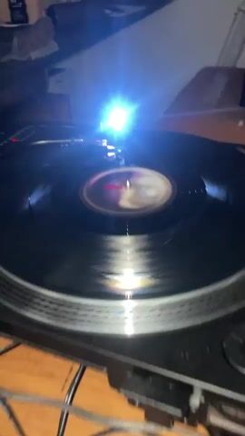 The video shows a turntable in operation. A black vinyl record spins with a colorful label. The needle rests on the record as it plays music. A bright light illuminates the scene, creating glare and obscuring some details. The turntable appears to be sitting on a surface with a tiled pattern visible at the bottom of the frame. In the background, there are glimpses of a counter or shelf holding various items, including a bottle. The camera remains mostly still, focusing on the spinning record and the needle.