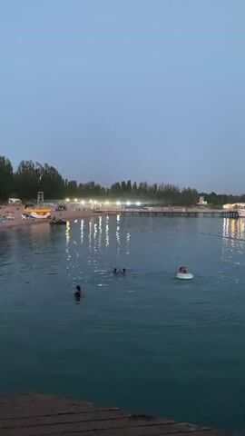 The video opens with a wide shot of a body of water, possibly a lake or sea, at dusk. The sky is a pale blue, transitioning to a darker shade of twilight. The water is calm, with a few ripples. In the distance, a shoreline is visible, lined with trees and a long pier structure. Lights are illuminated along the pier, reflecting on the water's surface. A white boat is docked near the pier. On the water, several people are swimming or playing. One person is on a white inflatable raft, and two others are swimming. Towards the foreground, a woman wearing a dark dress with a red and black pattern is visible from the back, standing on a wooden surface. As the video progresses, the camera pans slightly to the right, capturing more of the water and the distant shoreline. A fishing rod with a colorful line is visible in the lower right corner of the frame, extending into the water. The moon is faintly visible in the sky. The video seems to capture a serene evening scene at a waterfront location.