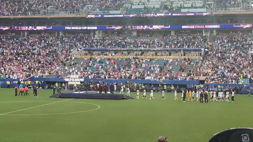 This is a video of a soccer stadium with a large crowd in the stands. On the field, players in uniform are lined up, some on the left and some on the right, facing a central platform. There are also people in suits and officials on the field. Banners overhead read "FIFA WORLD CUP 2026" and "BRONZE FINAL WINNERS." The overall scene suggests a medal ceremony or presentation after a soccer match.