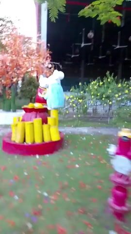 This video shows an outdoor scene, possibly a garden or park. In the center is a red, heart-shaped base adorned with several yellow cylindrical objects. Atop this base are two white mouse figurines, one dressed in red and the other in blue. They are holding a golden circular object between them. The ground is covered with green grass and scattered colorful petals. In the background, there are trees with some autumn-colored foliage and yellow flowers. A building structure with a dark roof and some lights is also visible behind the figurines. The camera pans slightly to the right, showing more of the surroundings.
