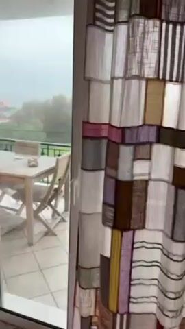 The video shows a balcony with a wooden table and chairs, and beyond the balcony railing is a view of trees, cars, and buildings. The weather appears to be rainy or foggy, reducing visibility. A patchwork curtain hangs partially over the sliding glass door that leads to the balcony. A hand briefly opens the curtain to reveal the outdoor scene.