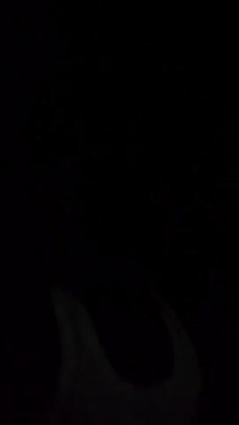 A person wearing a tank top stands in darkness. The video is shot in low light conditions, and the person is partially obscured. White light streaks are visible in the background, suggesting movement or a light source.