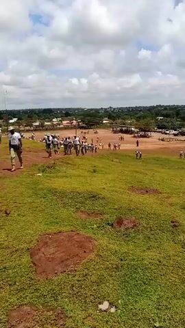 The video depicts a group of people running across a grassy field. Most of the people appear to be wearing white shirts and dark pants. A few are also wearing hats. The field is uneven, with patches of bare soil visible. In the background, there are trees and a few buildings. The sky is partly cloudy. The camera angle changes, and there is some shaking, suggesting the video was taken while moving. At least one person is wearing a shirt with “NYSC” visible.