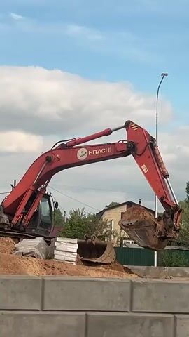 A red Hitachi excavator is shown in operation at a construction site. The excavator arm lifts a bucket of dirt and rotates to the right. A second excavator bucket sits in the dirt nearby. Piles of concrete blocks line the edge of the construction site, with grass and some houses visible in the background. The sky is blue with clouds.