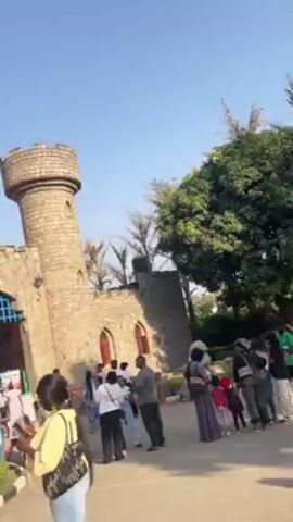 The video shows the entrance to Magic Land.  It is a castle-like structure with two towers on either side of the entrance. The words "Magic Land" are displayed in colorful letters above the gate. A Nigerian flag is visible on the left tower. People are gathered near the entrance, some are taking pictures. The sky is clear and blue.