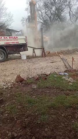 The video shows a drilling rig operating in a gravelly area with a truck parked nearby. The rig is tall and yellow, and it's drilling into the ground, creating a cloud of dust or vapor. The truck is white with red accents and has text in Bulgarian that translates to "Drilling for water, Drilling in rocks and mixed terrain," followed by a phone number. A white bucket sits on the back of the truck. The ground is covered with gravel and patches of grass. There are trees in the background, their branches bare. The sky is overcast, creating a hazy atmosphere. The camera is mostly static, providing a clear view of the drilling operation.