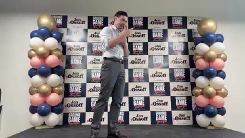 A man is standing on a stage or elevated platform, speaking into a microphone and gesturing with his right hand. He is wearing a light-colored button-up shirt, dark pants, and dark shoes. He has short dark hair and appears to be speaking passionately.

Behind him is a large banner with a checkered pattern. The banner repeatedly displays the words "GROUND GAME" and "Jon Ossoff Georgia" in black and white, with "Georgia" in a smaller red font. On either side of the banner are two tall balloon columns. These columns are made up of balloons in dark blue, white, gold, and light pink, with a large gold balloon at the very top of each column. The setting appears to be an indoor event, possibly a political rally or campaign stop in Decatur, United States, given the visible text on the banner.