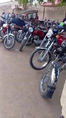 The video depicts an outdoor market scene. Numerous motorcycles are parked on an unpaved surface, with several men standing around them. Some men are dressed in light blue robes and caps, while others wear more casual attire. In the background, there are piles of grain and large white sacks on the ground, indicating agricultural goods for sale. Buildings with light-colored walls are visible in the distance, and trees provide some shade. The overall atmosphere suggests a bustling marketplace in a rural or semi-urban setting.