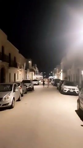 The video shows a nighttime street scene in a town. Buildings line both sides of the street, and cars are parked along the sides. There are streetlights illuminating the scene. In the distance, two figures, one appearing to be an adult and the other a child, are walking away from the camera. The video then pans to the side, showing details of the buildings, including awnings and windows. Finally, the video shows a sign with symbols, possibly a traffic sign, and then pans back up towards the dark night sky.