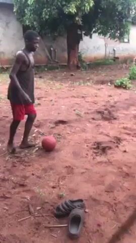 In a yard with a dirt ground, four young men play with a ball. One of the men wears red shorts and is shirtless. Another wears a striped shirt and dark shorts. One is in all black, and the fourth is wearing blue jeans and a dark jacket. A tree grows in the background, and a small building is behind them. A pair of sandals sit discarded on the ground near the bottom of the frame.