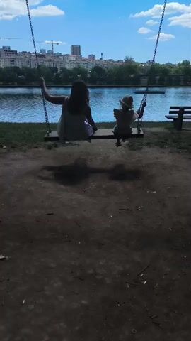 Two figures, one adult and one child, are seen on a swing set. The adult is on the left, and the child is on the right. They are both facing away from the camera, towards a body of water. In the background, there is a cityscape with buildings and trees. The sky is blue with some clouds. The swing set is in a park area with a dirt ground. The adult appears to be wearing a white dress, and the child is wearing a hat and a light-colored outfit. The adult is holding onto one of the swing chains, and the child is also holding onto the chains. They are both gently swinging back and forth.