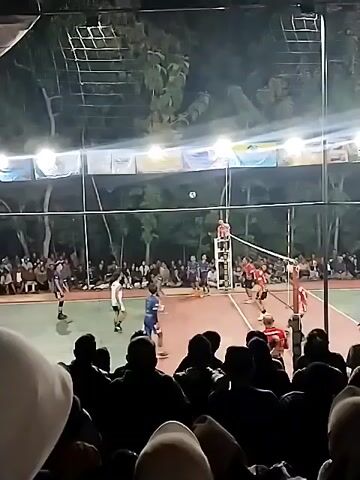 A night-time volleyball game is being played on an outdoor court illuminated by overhead lights. A crowd is gathered to watch the match, positioned in the foreground and on the sidelines. Two teams, distinguishable by their uniforms (one in red and white, the other in blue and white), are actively engaged in the game. The ball is in play, and players are positioned on both sides of the net, with some in the process of serving, receiving, or setting. The court surface appears to be a reddish-brown color, and the surrounding environment is dark with trees visible in the background. The spectators are largely silhouetted against the court lights, making their individual features indistinct. The atmosphere seems lively, typical of a sporting event.