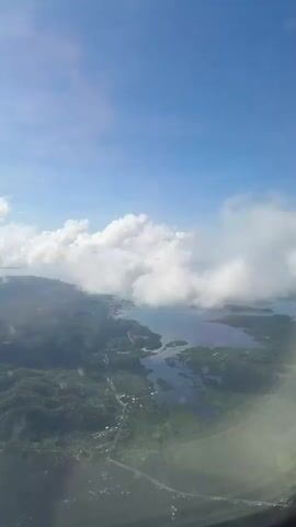 The video shows an aerial view from an airplane window. Below, there's a landscape of green forest and islands surrounded by water. A road is visible winding through the trees. Large, fluffy white clouds are scattered in the sky, partially obscuring the view. The sky is a clear, bright blue. The airplane window reflects some interior elements.
