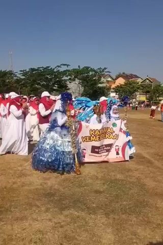 A group of people, mostly women in white robes and red headscarves, are participating in an outdoor event. They are walking and clapping. Two individuals in elaborate blue and white costumes, resembling large dresses, are standing prominently in the center. One of these costumes appears to be made from plastic bottles. A banner with text and images is visible behind one of the costumed individuals. The event seems to be taking place on a dry grassy field under a clear blue sky, with houses and trees in the background. There are also other people and structures, possibly stalls or tents, further in the distance.