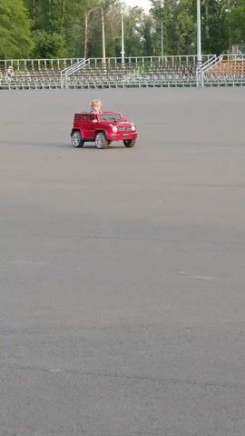 A red toy Mercedes G-Wagon with a young blonde child inside is driving on a paved surface. The car is moving from left to right. In the background, there are bleachers and trees. The lighting suggests it is daytime.
