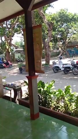 The video shows an outdoor scene with a paved area in the foreground and background. In the foreground, there is a planter box filled with green plants. Behind the planter box, there are several motorcycles parked. A white scooter is on the far left, followed by two red-and-black motorcycles. A dark motorcycle is parked further to the right. Beyond the motorcycles, there are trees and some buildings with red roofs. A banner with text is hanging on a pole on the left side of the image. The lighting suggests it is daytime.